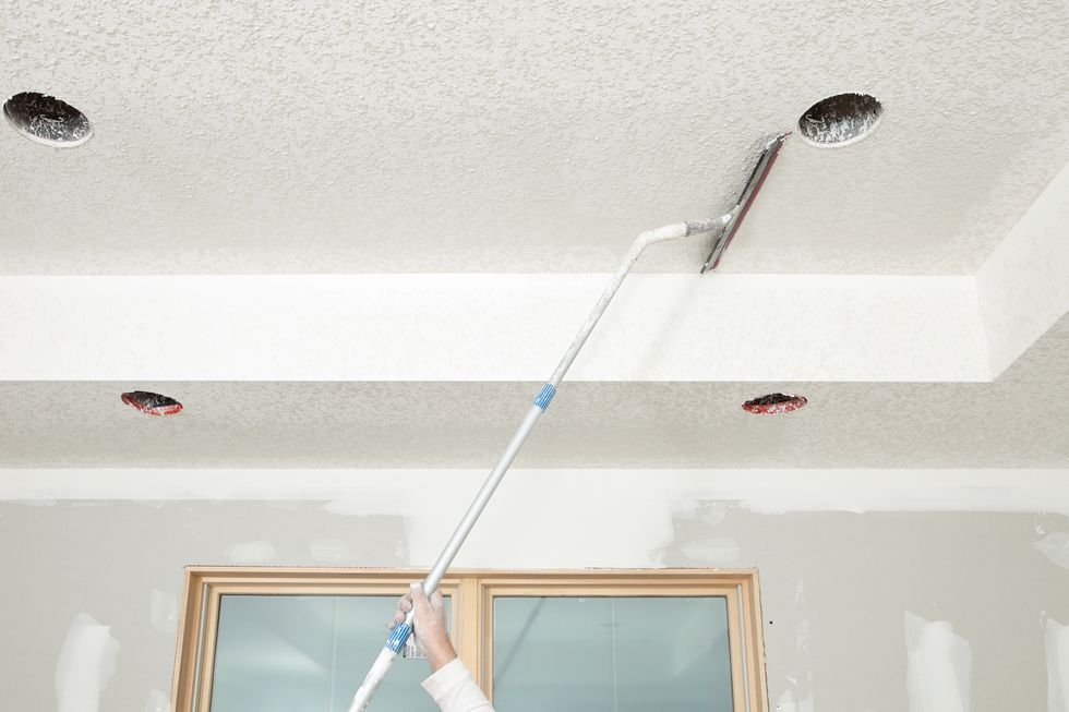 Một công nhân vách thạch cao đang trát trên bùn vách thạch cao đã được phun, bằng lưỡi cao su, để tạo họa tiết hạ trần phần trên của hình ảnh vẫn chưa được làm phẳng, phần khung giữa đã hoàn thiện